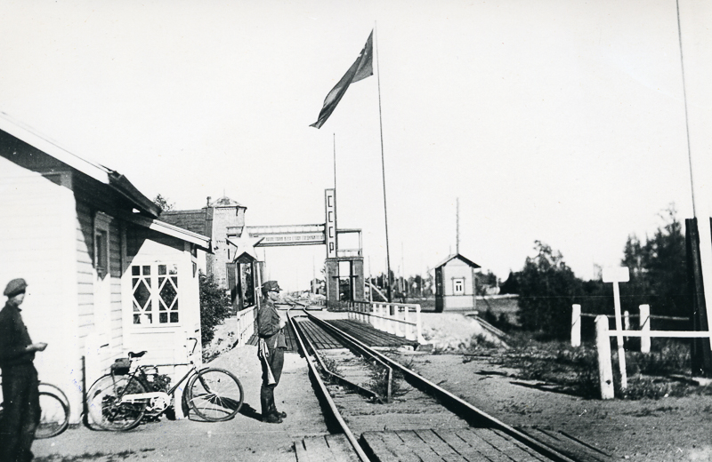 Raide ja rautatiesilta Suomen ja Neuvostoliiton rajalla. Raiteen yllä näkyy suuri tähti ja teksti CCCP. Lipputangossa liehuu lippu ja raiteen vieressä seisoo sotilas. Vasemmalla seisoo toinen ihminen.