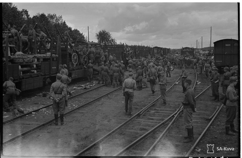 Raiteilla siesoo junavaunuja johon on lastattu kärryjä. Vaunujen kuormatiloissa ja ympärillä on paljon sotilaita. Kauempana junien lähellä on hevosen vetämät kärryt.