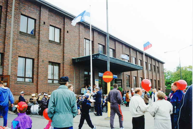 Ihmisiä asemarakennuksen edessä. Vasemmalla näkyy musiikkiyhtye ja rummut. Rakennuksen edessä on aksi lipputankoa, toisessa on Suomen lippu ja toisessa Venäjän lippu.
