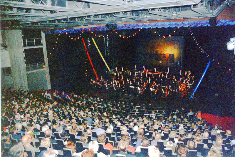 Konserttisali, jonka valoilla koristellulla lavalla on orkesteri ja katsomossa yleisöä.