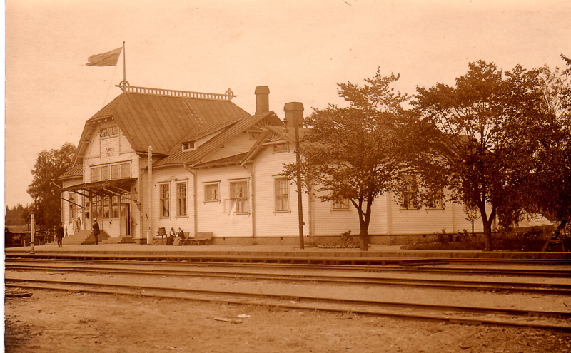 Vanha asemarakennus, jonka lipputangossa liehuu lippu. Rakennuksen edustalla asemalaiturilla seisoo ja istuu ihmisiä. Laiturin edessä on raiteita. Asemarakennuksen vieressä on puita.