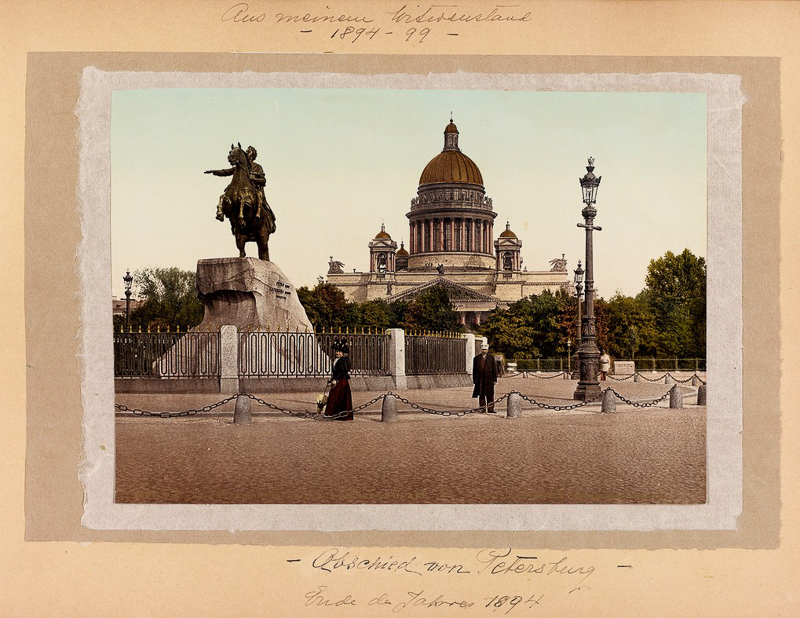 Postikortti Pietarista, jossa on kuvattu ratsastajapatsas Vaskiratsastaja. Taustalla oleva kirkko on Iisakinkirkko. Etualalla on mukulakivinen katu ja kaksi ihmistä.