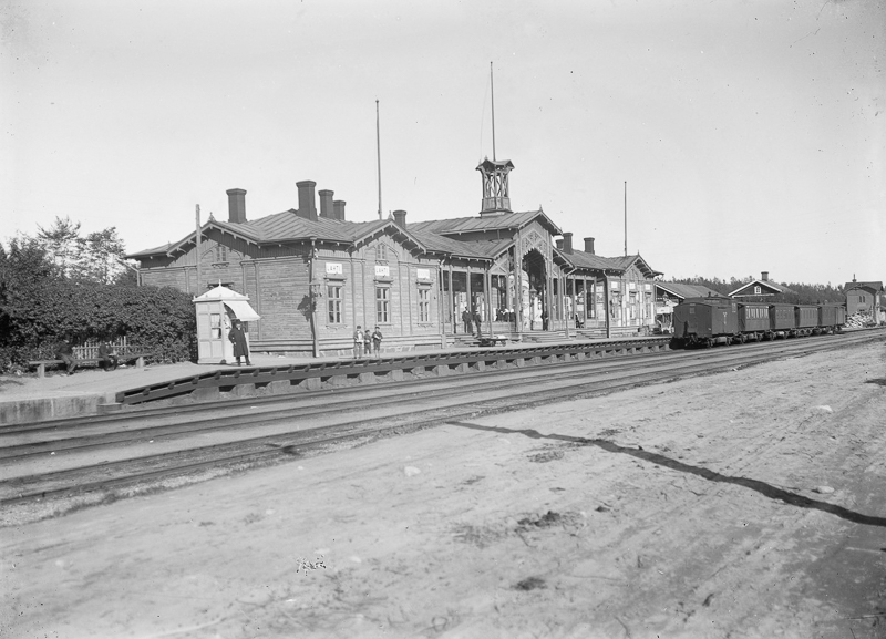 Asemarakennus, jonka edessä on junaraiteet. Raiteilla seisoo vaunuja. Asemalaiturilla on muutamia ihmisiä. Aseman oikealla puolella taustalla on rakennuksia.